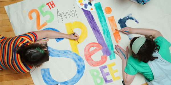 3 students lay on the floor of a gym coloring a poster for the 25th annual spelling bee