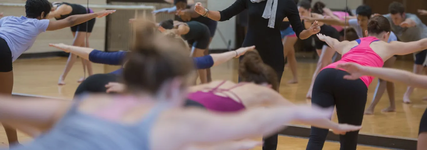 A woman wearing a black body suit and scarf, leads a crowded room of dancers